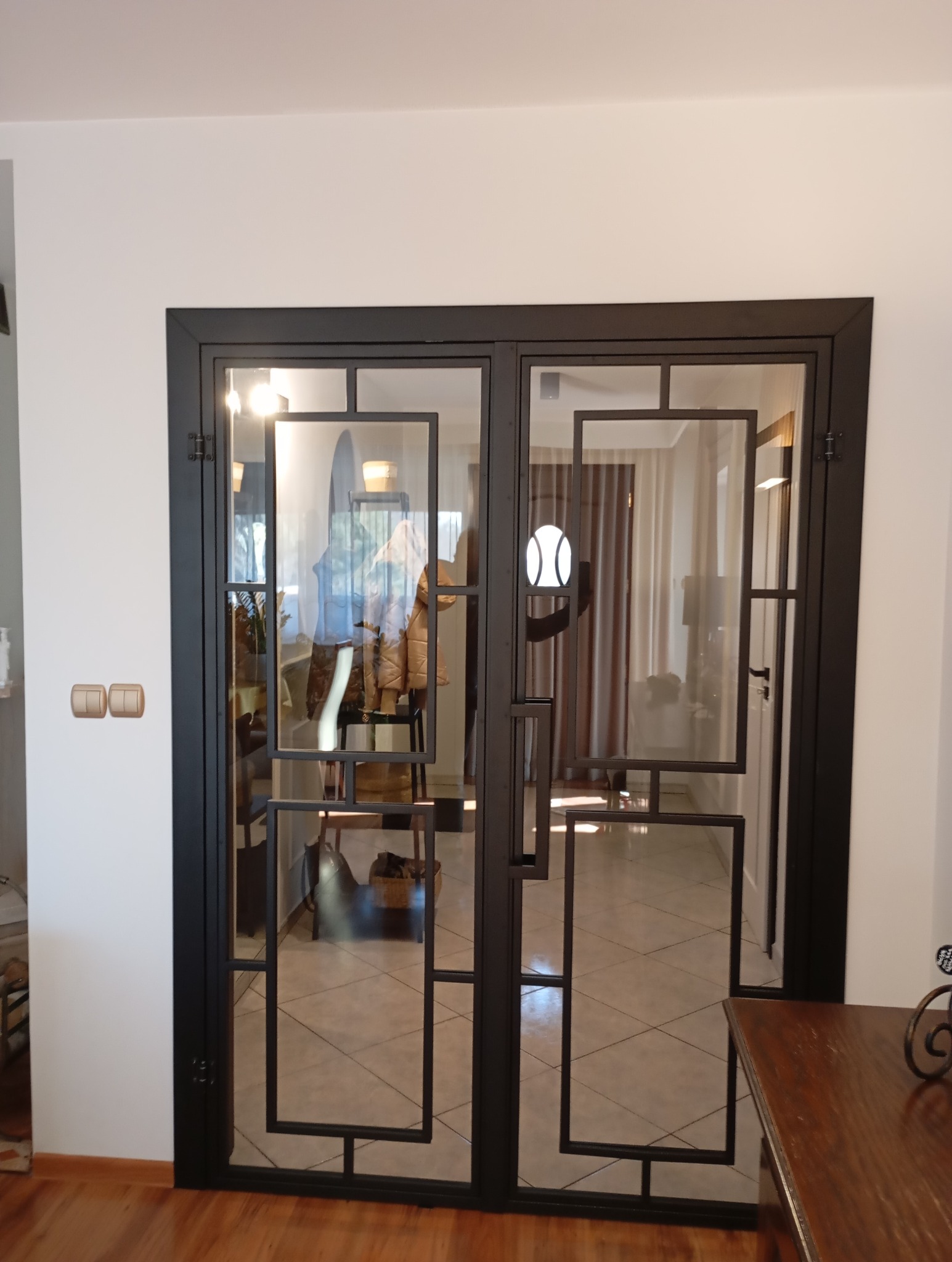 Dwuskrzydłowe drzwi wewnętrzne z czarną, metalową ramą i szklanymi panelami, wykonane w Przybyszewie, z geometrycznym wzorem. Widoczne odbicie wnętrza domu.