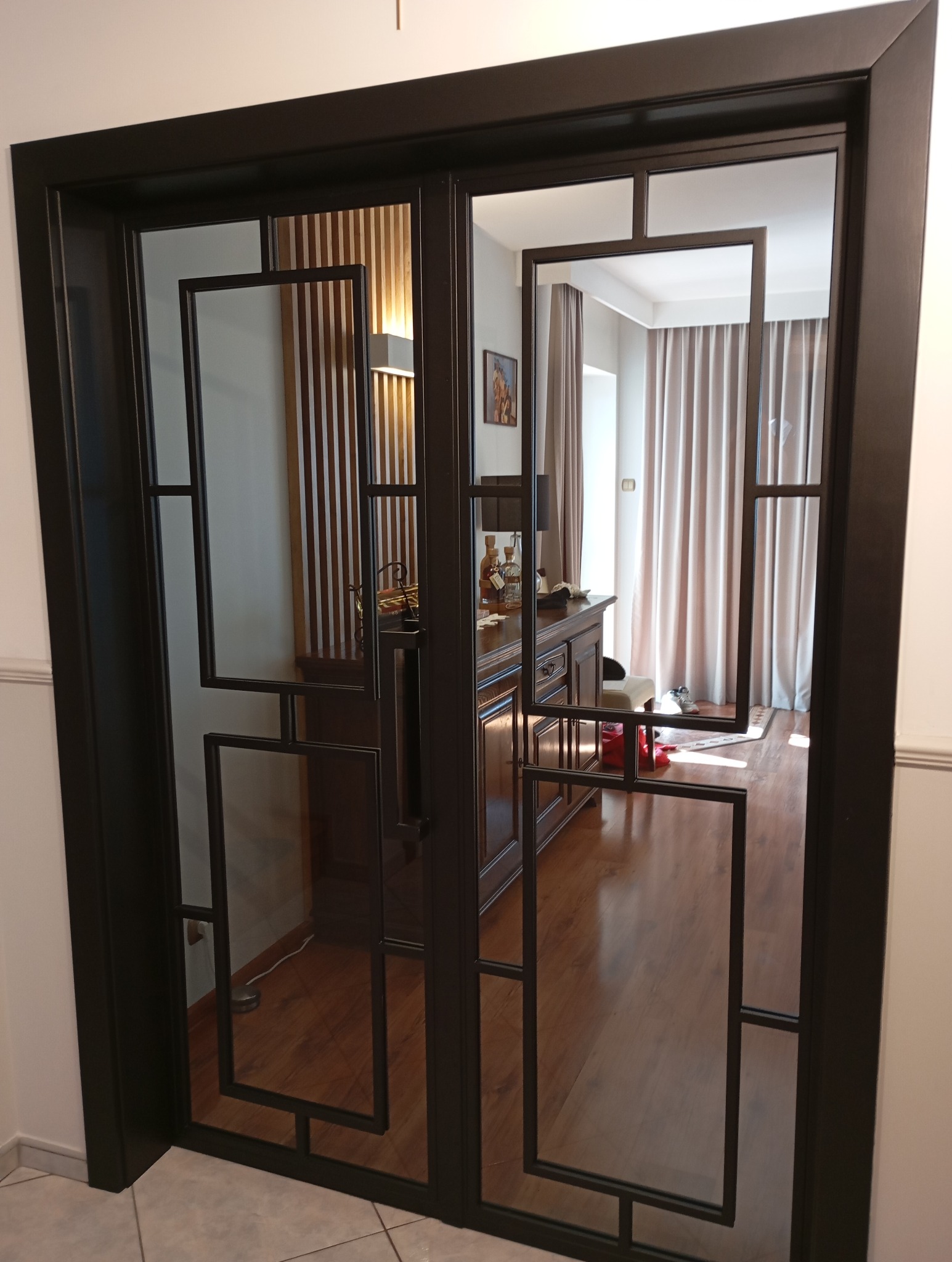 Dwuskrzydłowe drzwi wewnętrzne z czarną, metalową ramą i geometrycznymi podziałami szklanymi, odbijające wnętrze salonu z drewnianą komodą i zasłonami.