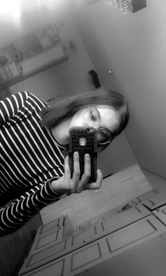 Osoba w okularach i bluzce w paski robi selfie w lustrze łazienkowym z widoczną szafką pod umywalką.