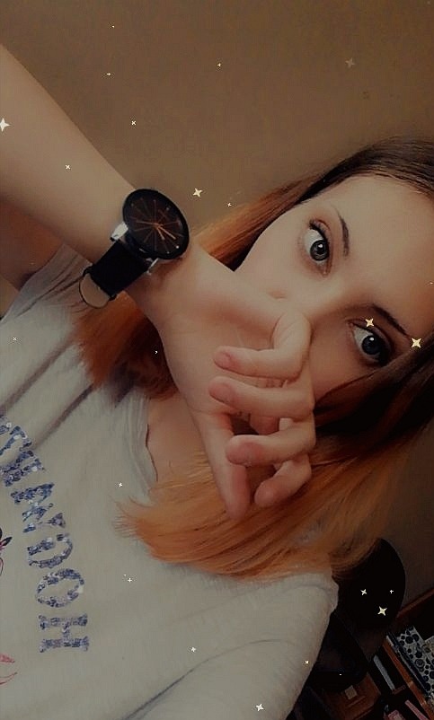 Młoda kobieta z pomarańczowymi końcówkami włosów, zakrywa usta dłonią, na nadgarstku czarny zegarek na skórzanym pasku, na bluzce błyszczący napis