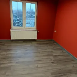 Pusty pokój z pomalowanymi na czerwono ścianami, panelami podłogowymi w odcieniach szarości i białym grzejnikiem pod oknem z widokiem na zimowy krajobraz.