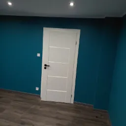 Świeżo wykończone wnętrze z turkusowymi ścianami, białą, nowoczesną stolarką drzwiową i podłogą imitującą ciemne drewno, oświetlone punktowo.