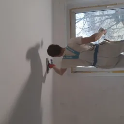 Pracownik w kombinezonie Knauf szpachluje ścianę w pobliżu okna, używając pacy i rękawic ochronnych.