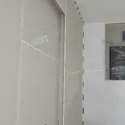 Wnętrze w trakcie montażu płyt kartonowo-gipsowych, widoczne łączenia płyt wzmocnione taśmą siatkową, otwór na instalację elektryczną i częściowo zasłonięte okno folią ochronną.