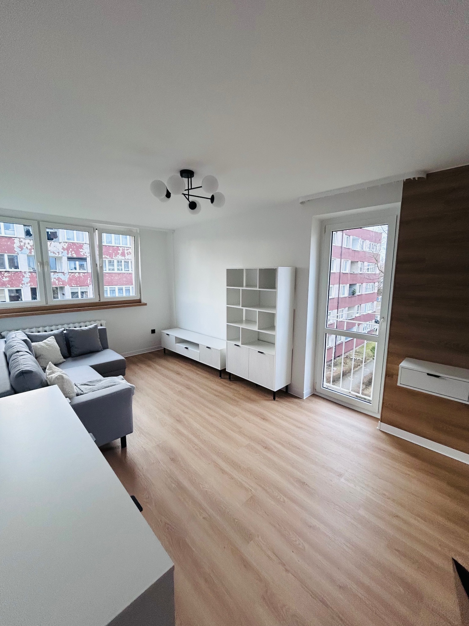 Jasne wnętrze po remoncie: parkiet, szara sofa z poduszkami, białe meble, okno z widokiem na blok. Nowoczesna lampa sufitowa.