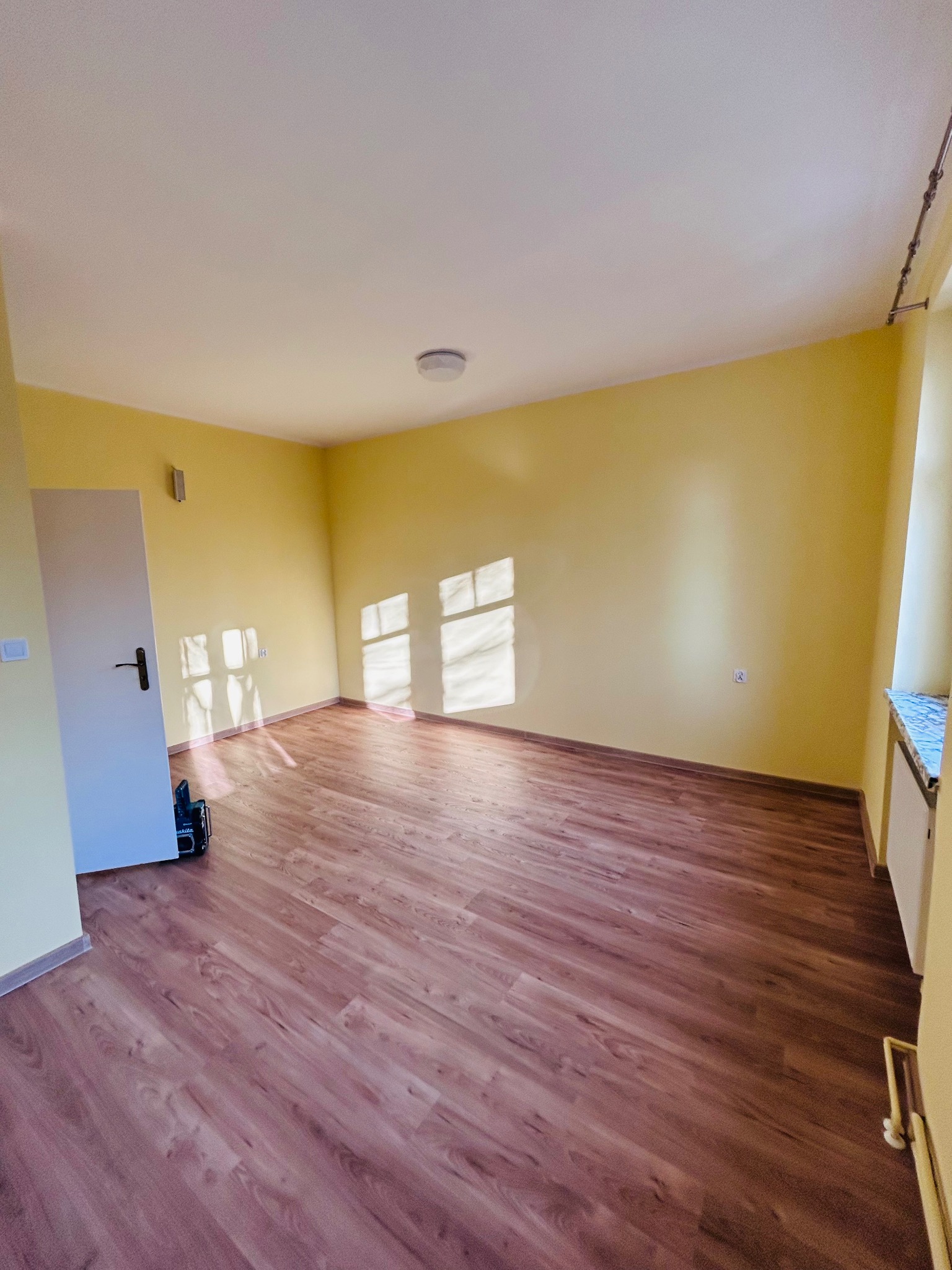 Jasne, odświeżone wnętrze pokoju po remoncie: żółte ściany, brązowe panele podłogowe, białe drzwi i okno z marmurowym parapetem. W rogu widoczny sprzęt.