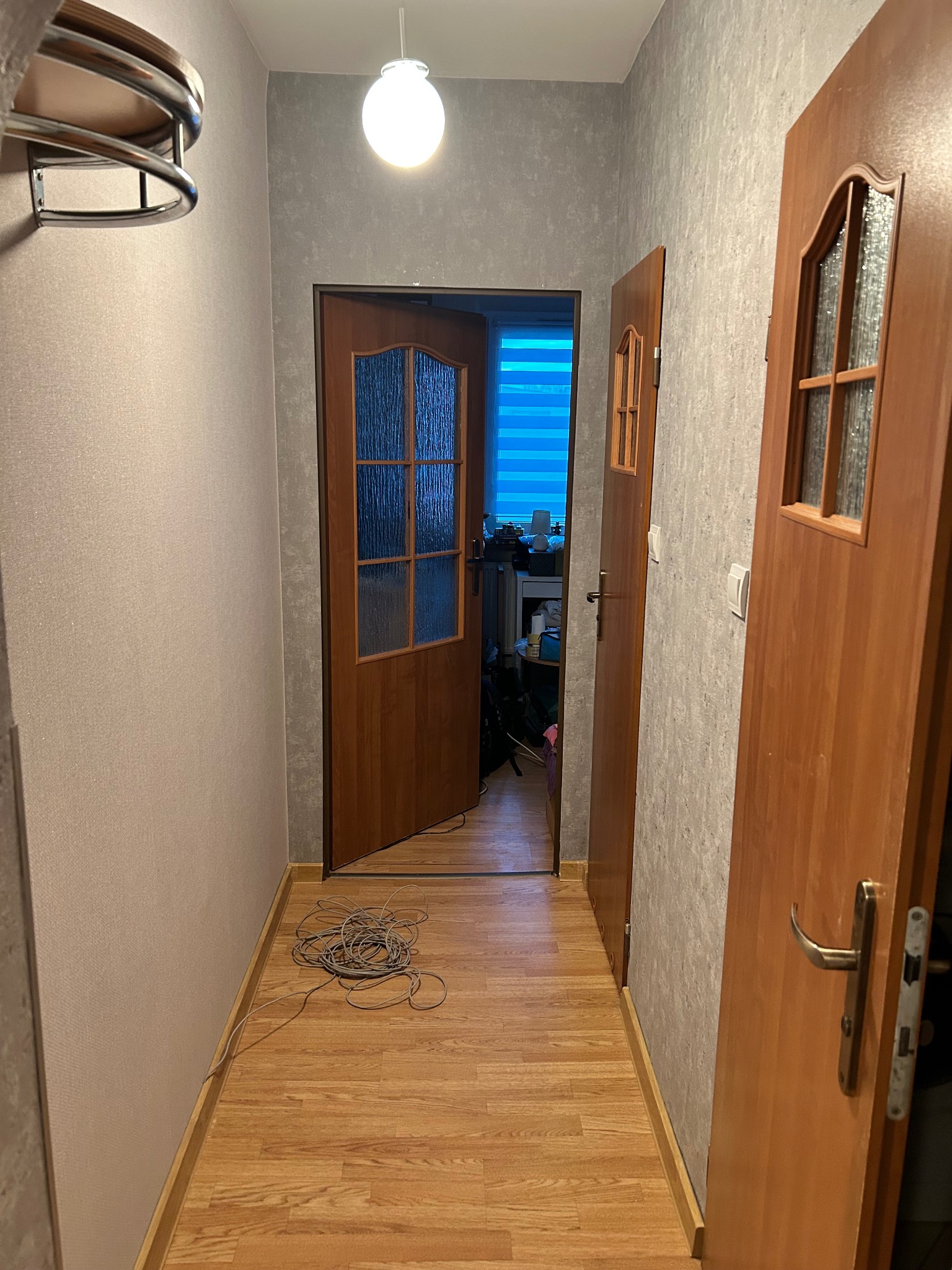 Korytarz po remoncie: świeżo położone panele podłogowe, ściany z nową tapetą o fakturze betonu. Widoczne dwie drewniane drzwi z przeszkleniami i zwisający kabel.