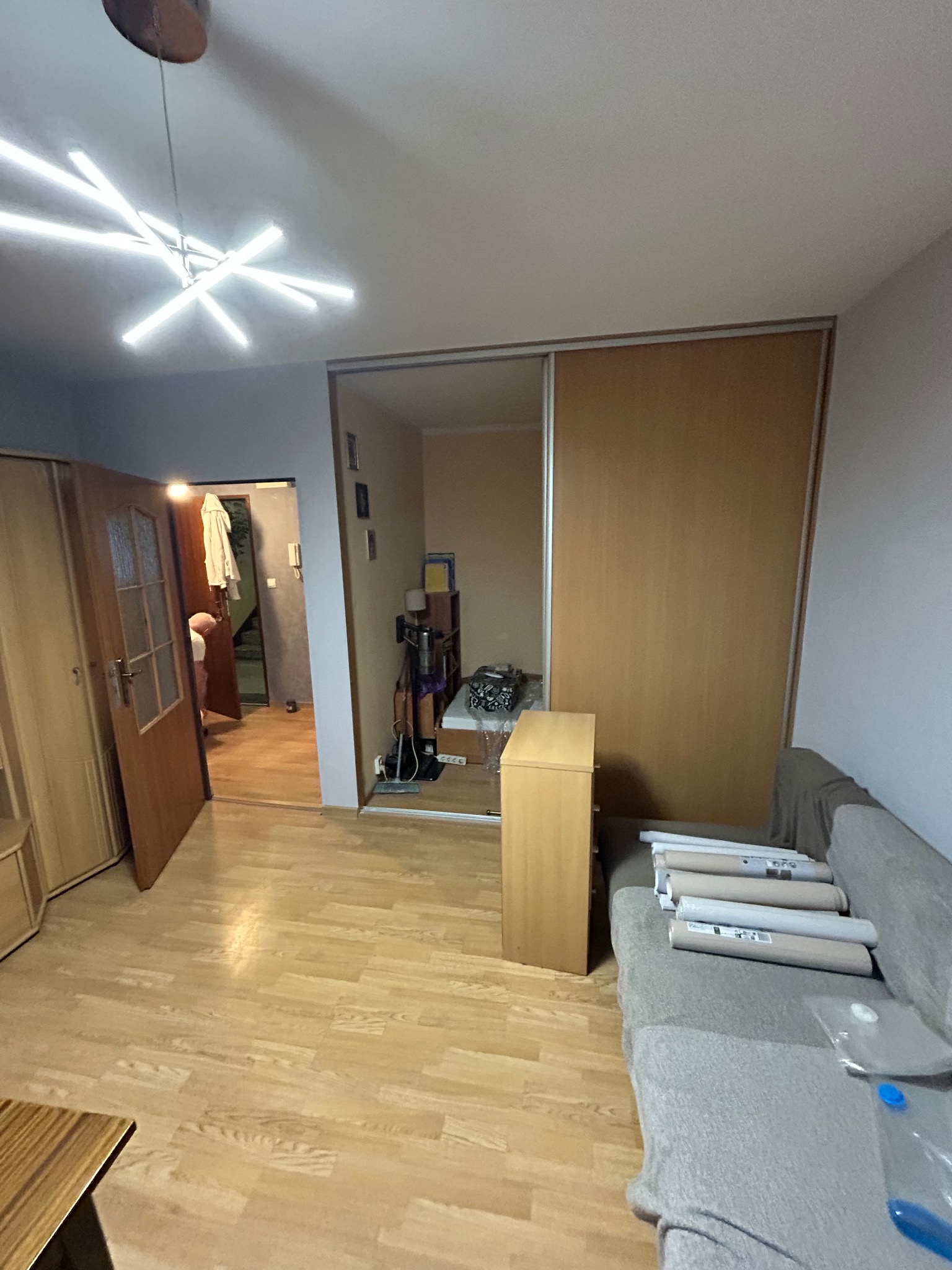 Jasne wnętrze pokoju z drewnianą podłogą, przesuwną szafą i nowoczesną lampą sufitową. Na kanapie leżą zwinięte plakaty lub tapety, sugerujące prace wykończeniowe.