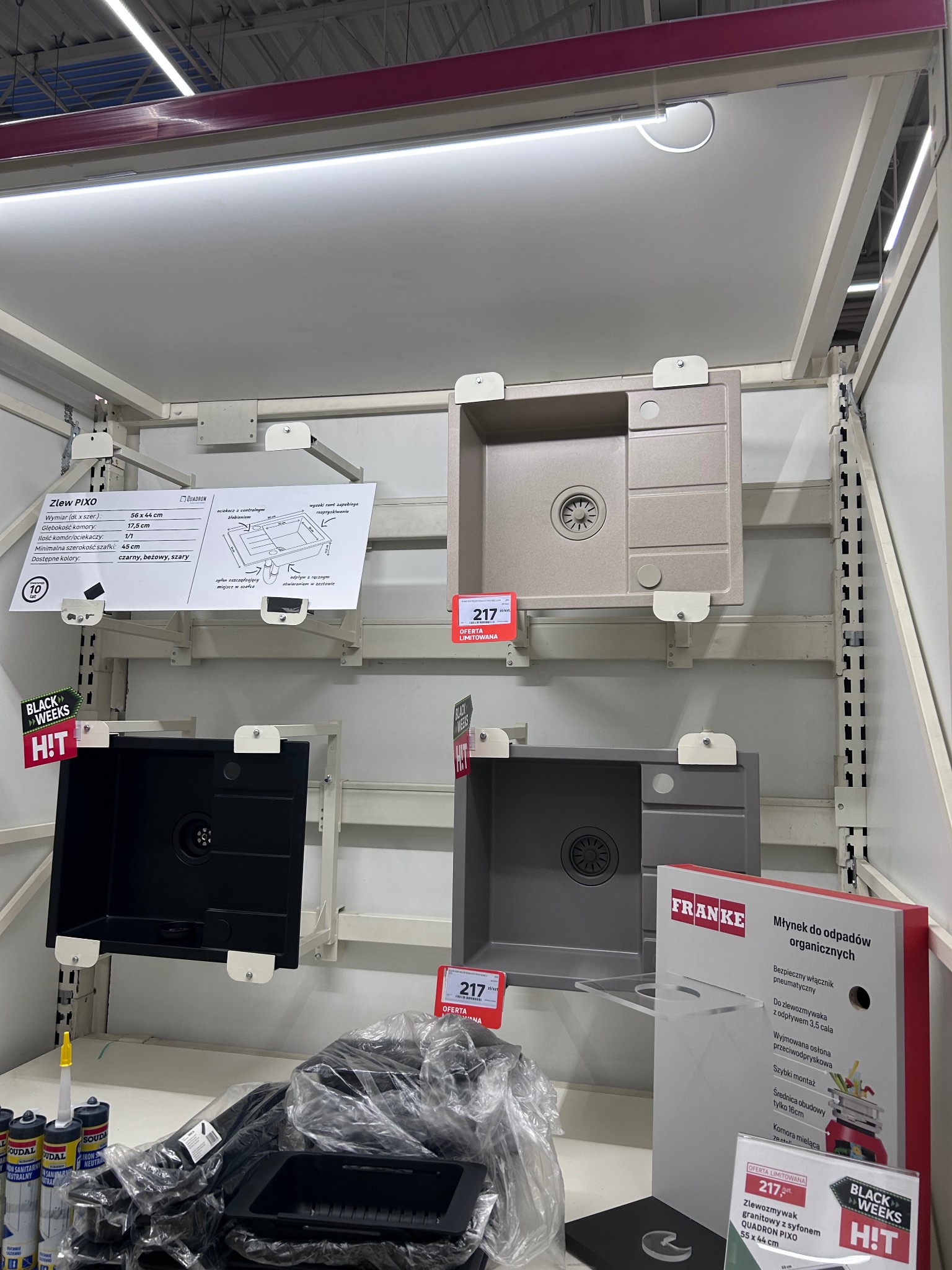 Ekspozycja zlewów granitowych w sklepie: czarny, beżowy i szary model Quadron PIXO z ceną 217 zł, obok młynek do odpadów Franke i akcesoria montażowe.