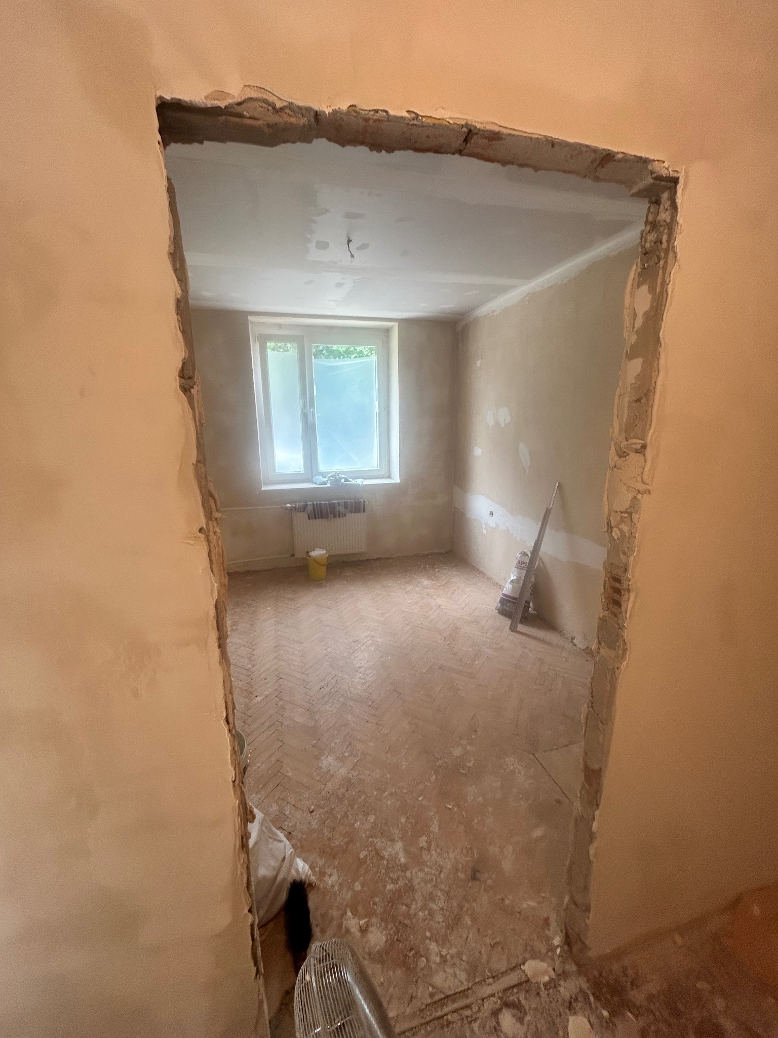 Wnętrze w trakcie remontu: widok na pokój przez otwór w ścianie, ściany częściowo otynkowane, podłoga z parkietu, okno z widokiem na zieleń. Etap prac wykończeniowych.