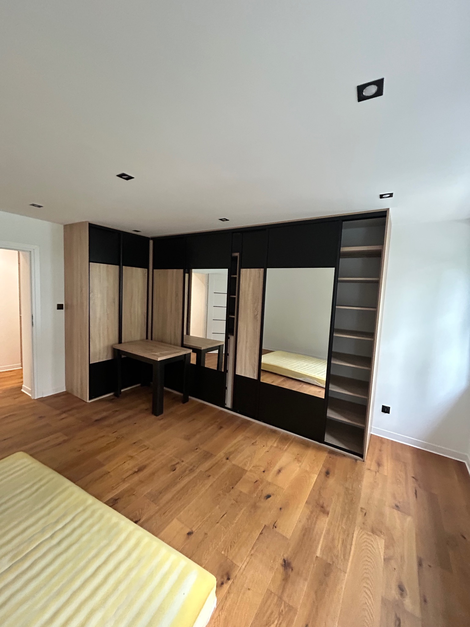 Nowoczesna szafa wnękowa z lustrami i drewnianymi elementami, w jasnym pokoju z parkietem. Widoczne oświetlenie sufitowe i minimalistyczny stolik.