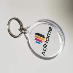 Okrągły, przezroczysty brelok z logo AdiHome, zawierający kolorowe paski w odcieniach czerni, żółci, czerwieni i błękitu.