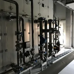 Kompleksowa instalacja grzewcza z rurami, zaworami i czujnikami, zamontowana przy szarej ścianie w hali przemysłowej, widoczne elementy mocujące i koryto kablowe.