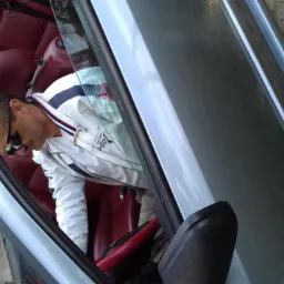 Mężczyzna w okularach wsiada do srebrnego kabrioletu z czerwoną skórzaną tapicerką. Ujęcie z boku, widoczne detale wnętrza i fragment otoczenia.