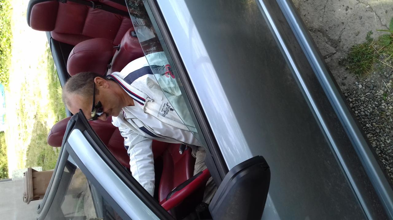 Mężczyzna w okularach wsiada do srebrnego kabrioletu z czerwoną skórzaną tapicerką. Ujęcie z boku, widoczne detale wnętrza i fragment otoczenia.