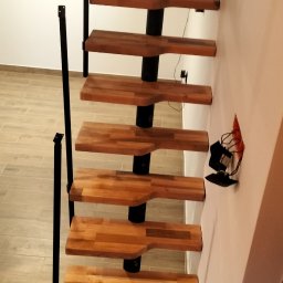 Artex - Spiralne schody z drewnianymi stopniami i czarną metalową konstrukcją, widok z dołu, z widoczną instalacją elektryczną w ścianie.