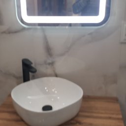 Artex - Nowoczesna łazienka z umywalką nablatową w kształcie zaokrąglonego kwadratu, czarną armaturą i szafką z lamelami na tle marmurowej ściany, w lustrze odbija się osoba robiąca zdjęcie.