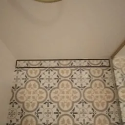 Fragment łazienki z ułożonymi dekoracyjnymi płytkami ceramicznymi w odcieniach beżu, szarości i bieli, z motywem roślinnym i geometrycznym, widoczny fragment złotej rury pod sufitem.