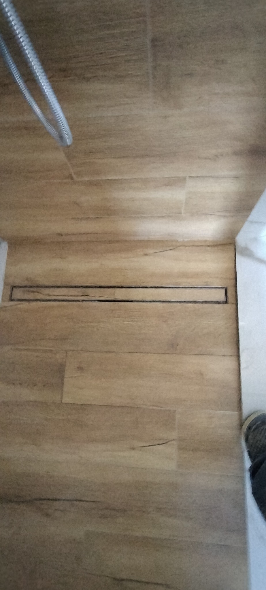 Ułożone płytki podłogowe imitujące drewno w łazience z odpływem liniowym, widoczne fragment węża prysznicowego i buta.