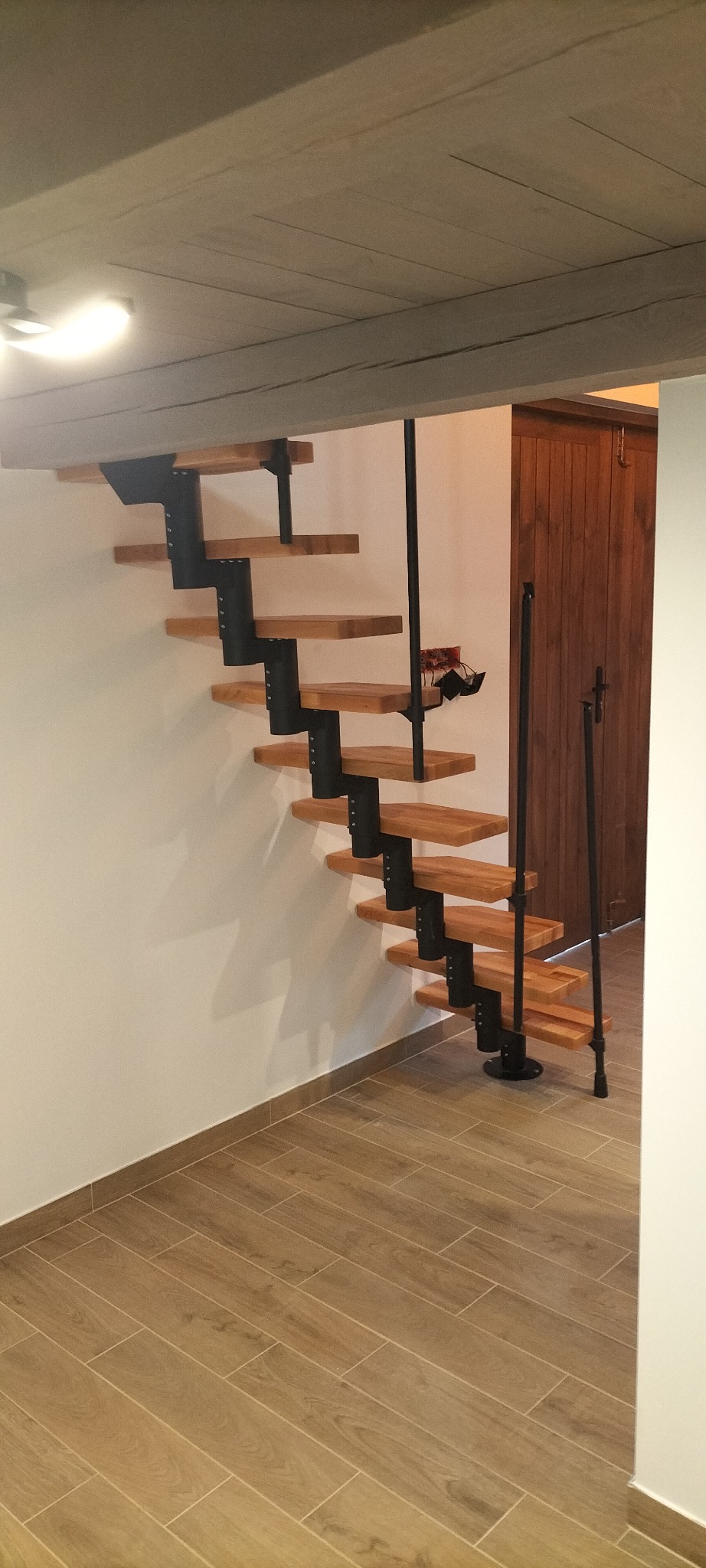 Nowoczesne schody ażurowe z drewnianymi stopniami i metalową konstrukcją, zamontowane przy białej ścianie, widoczne płytki podłogowe imitujące drewno i drewniane drzwi w tle.