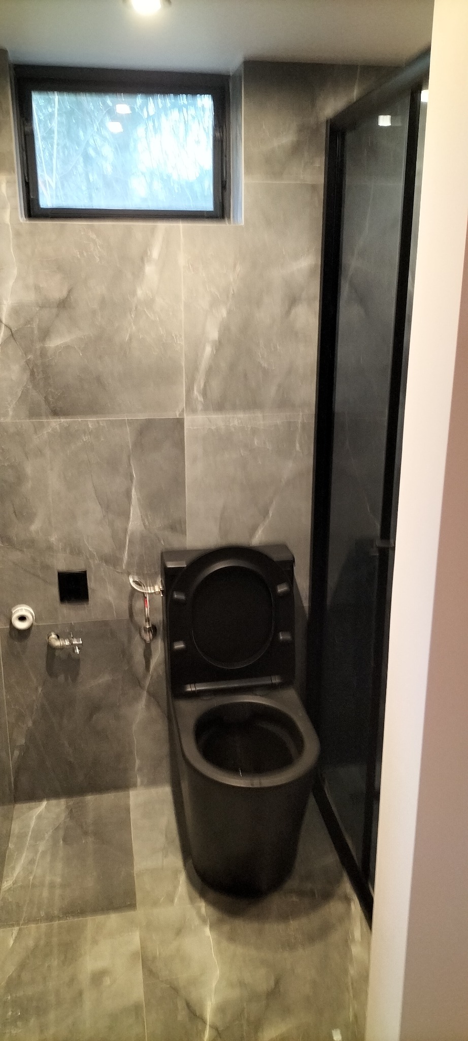 Nowoczesna łazienka z szarymi marmurowymi płytkami, czarną miską WC z otwartą klapą i kabiną prysznicową z czarną ramą.