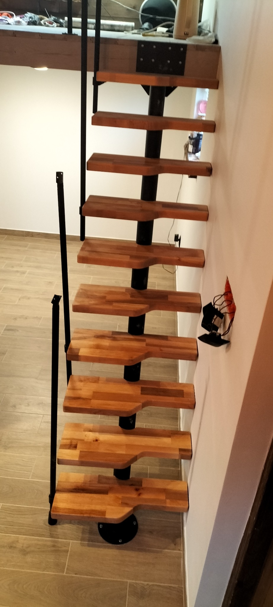 Spiralne schody z drewnianymi stopniami i czarną metalową konstrukcją, widok z dołu, z widoczną instalacją elektryczną w ścianie.