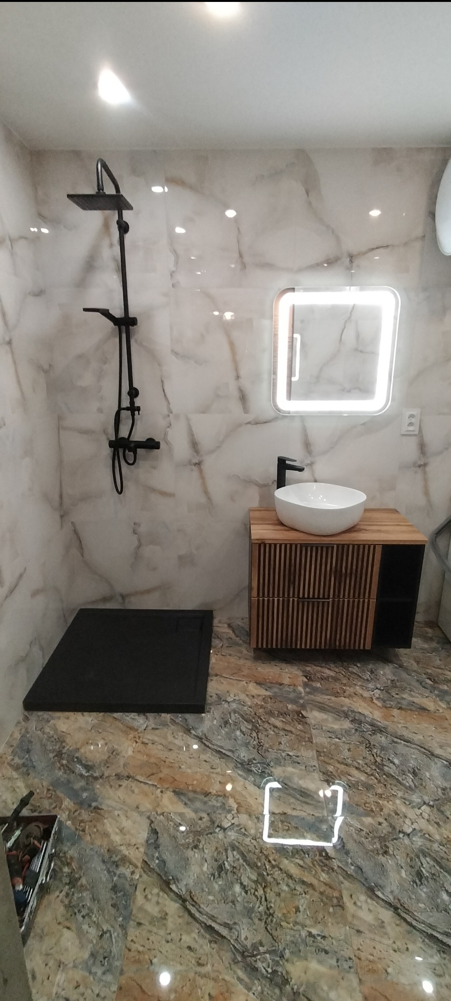 Nowoczesna łazienka z marmurowymi płytkami, czarną armaturą prysznicową i umywalką nablatową na szafce z drewnianymi lamelami oraz podświetlanym lustrem.