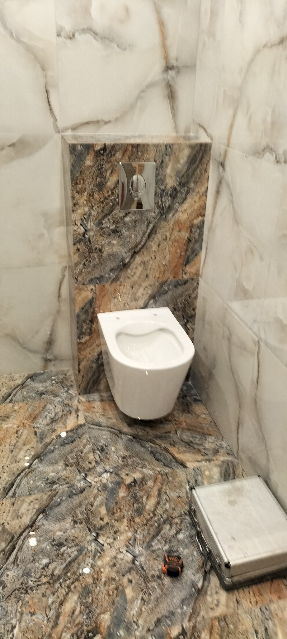 Nowoczesna toaleta podwieszana z chromowanym przyciskiem spłukiwania, obudowana płytkami imitującymi marmur w odcieniach bieli, beżu i brązu, z widoczną skrzynką narzędziową i miarką na podłodze.