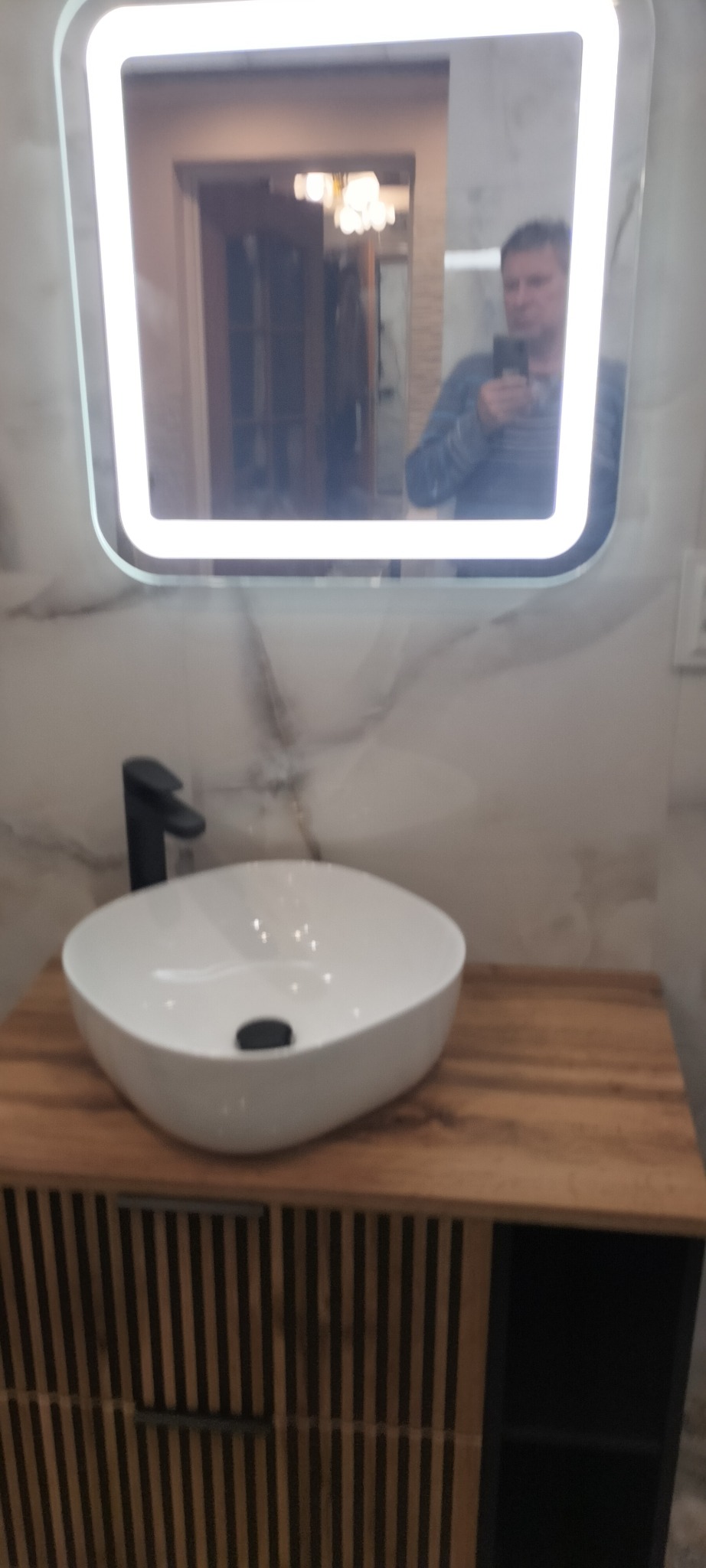 Nowoczesna łazienka z umywalką nablatową w kształcie zaokrąglonego kwadratu, czarną armaturą i szafką z lamelami na tle marmurowej ściany, w lustrze odbija się osoba robiąca zdjęcie.