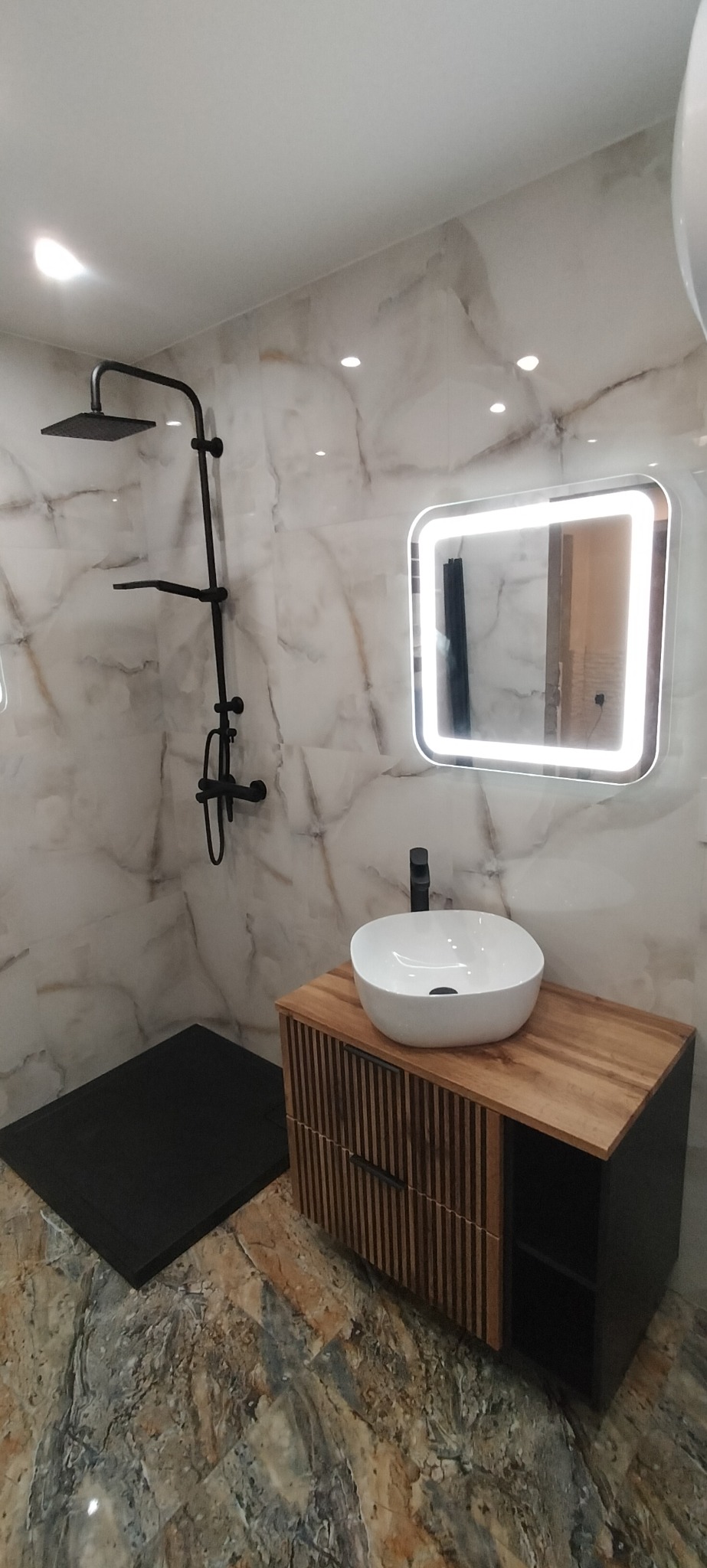 Nowoczesna łazienka z marmurowymi płytkami, czarną armaturą i podświetlanym lustrem nad umywalką nablatową na szafce z drewnianymi lamelami.