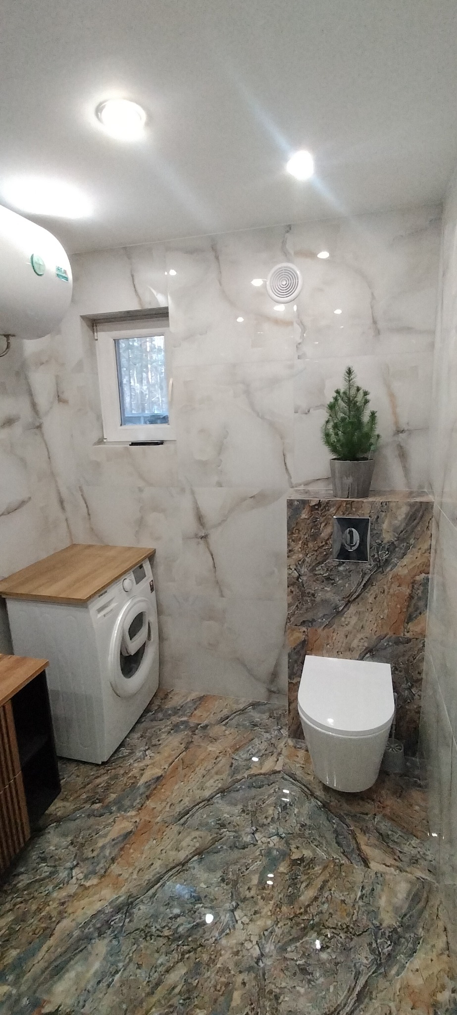 Nowoczesna łazienka wykończona marmurowymi płytkami, z pralką zabudowaną drewnianym blatem, wiszącą toaletą i małą choinką na postumencie.