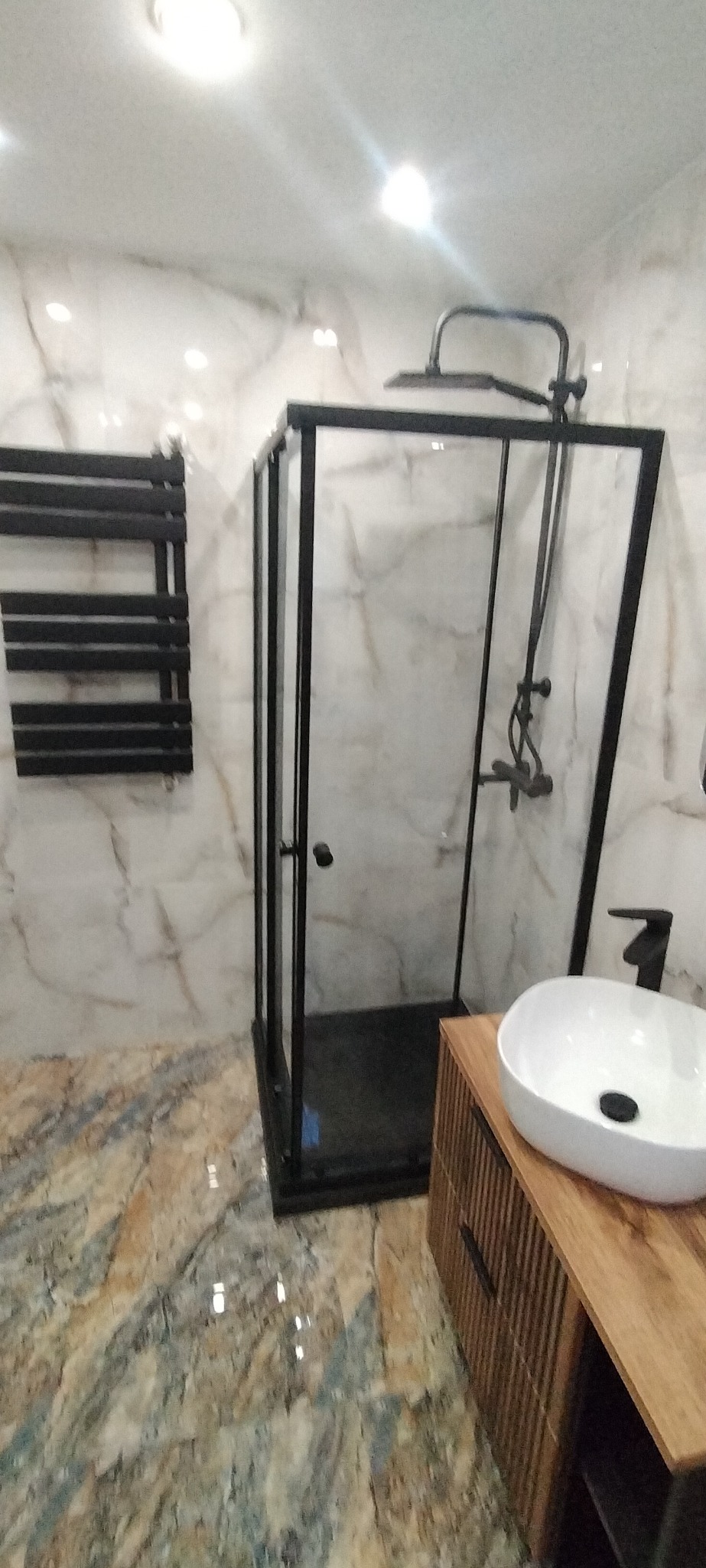 Nowoczesna łazienka z kabiną prysznicową typu walk-in z czarnymi profilami, wiszącym grzejnikiem drabinkowym i umywalką nablatową na szafce z drewnianymi lamelami.