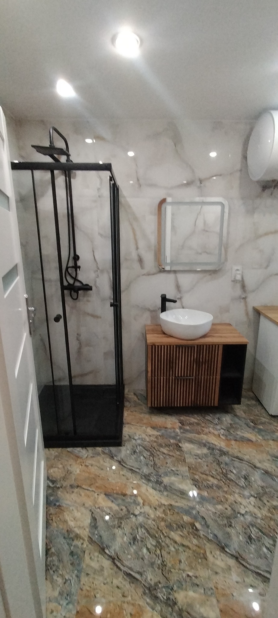 Nowoczesna łazienka z kabiną prysznicową typu walk-in z czarnymi profilami, umywalką nablatową na szafce z drewnianymi lamelami i podświetlanym lustrem, wykończona płytkami imitującymi marmur.