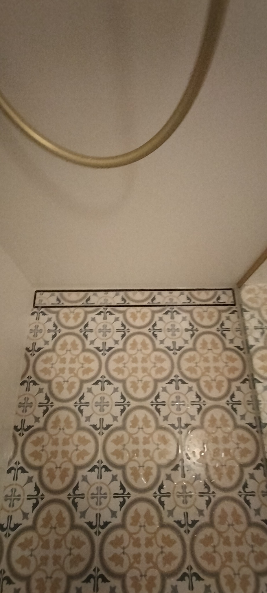 Fragment łazienki z ułożonymi dekoracyjnymi płytkami ceramicznymi w odcieniach beżu, szarości i bieli, z motywem roślinnym i geometrycznym, widoczny fragment złotej rury pod sufitem.