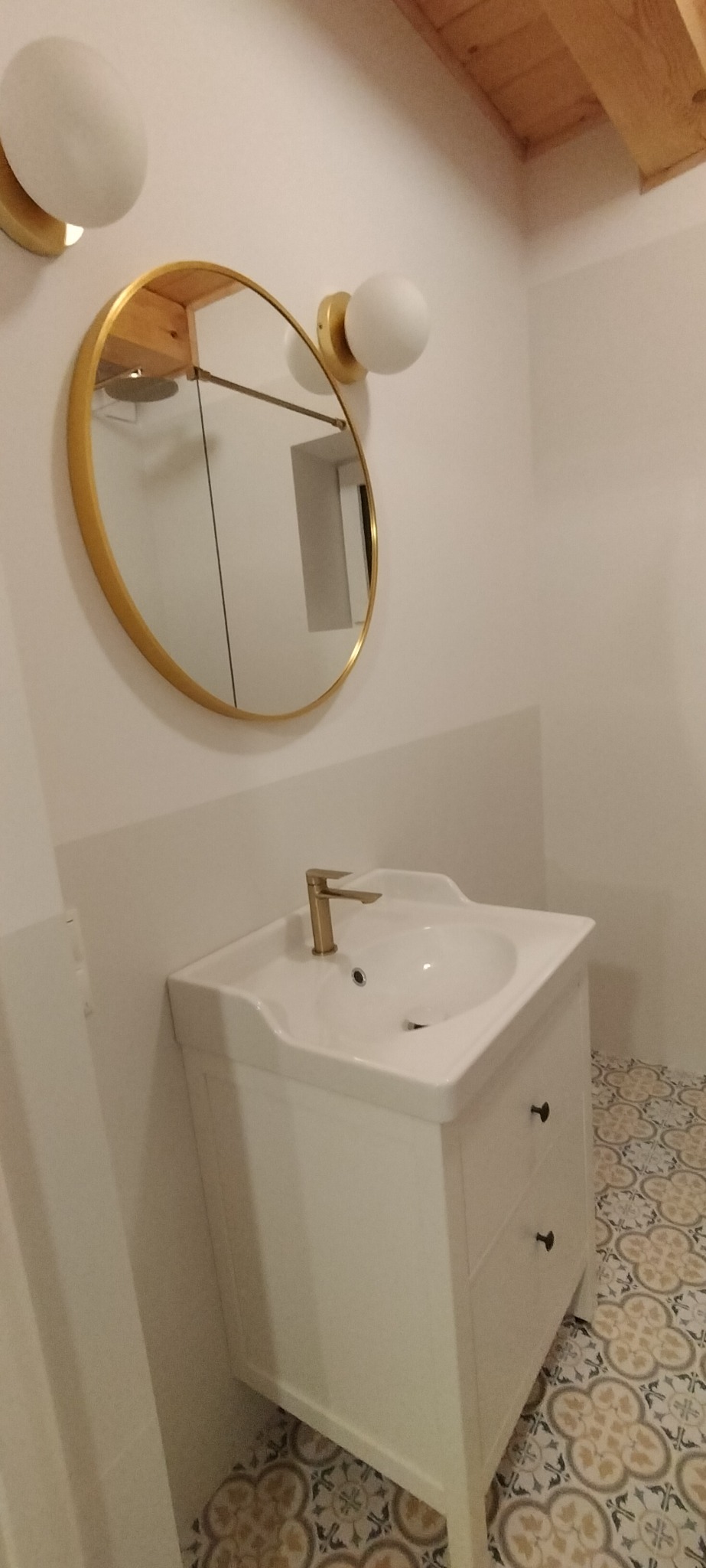 Biała szafka umywalkowa z dwiema szufladami i złotą baterią, okrągłe lustro w złotej ramie i dwie lampy-kule w łazience z wzorzystą podłogą.