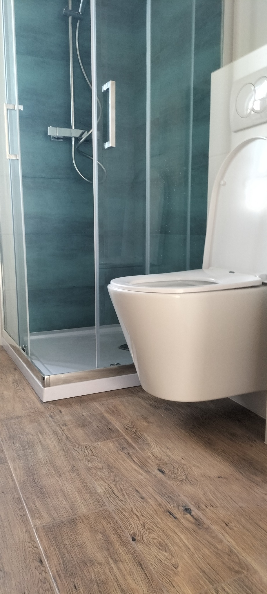 Nowoczesna łazienka po remoncie: kabina prysznicowa z turkusowymi płytkami i podwieszana toaleta na tle drewnopodobnych paneli podłogowych.