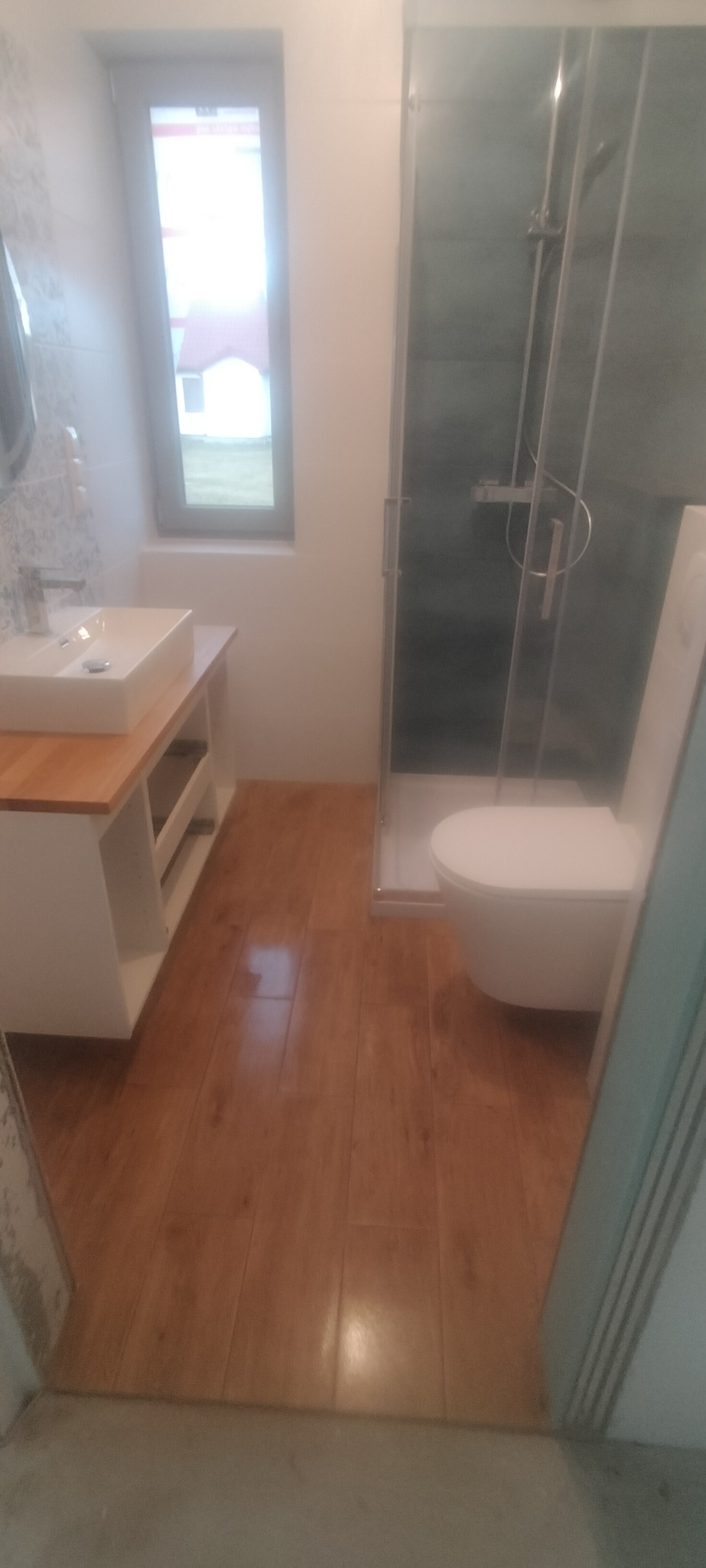 Nowoczesna łazienka z drewnopodobnymi panelami podłogowymi, białą umywalką nablatową na szafce z drewnianym blatem, kabiną prysznicową z szarymi płytkami i białą toaletą.