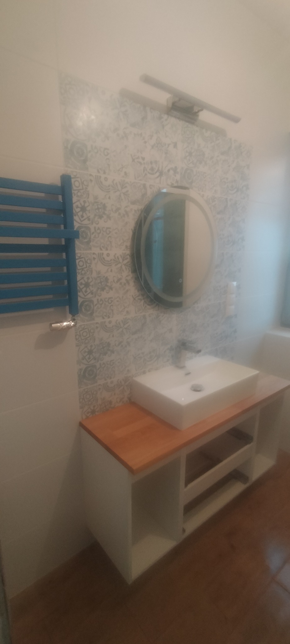 Nowoczesna łazienka z białą umywalką nablatową na szafce z drewnianym blatem, okrągłe lustro z podświetleniem, niebieski grzejnik drabinkowy i płytki w stylu retro.