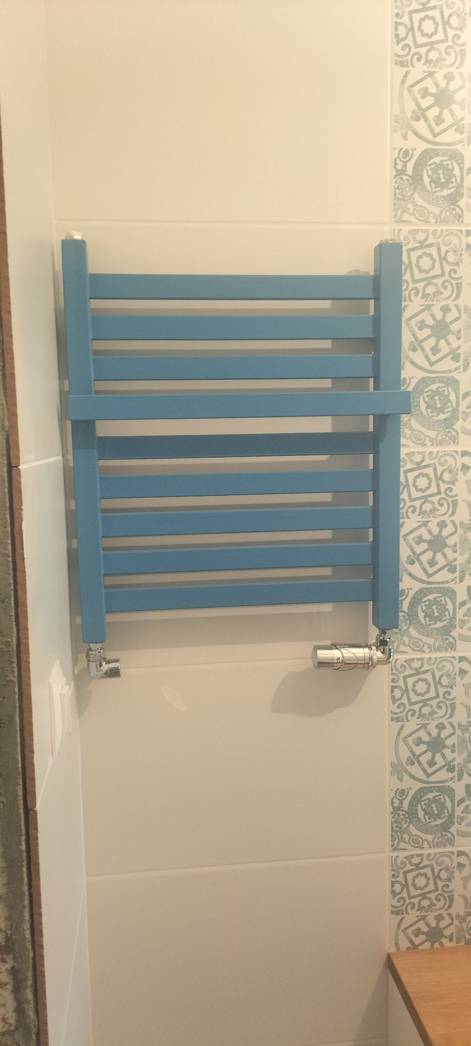 Niebieski, drabinkowy grzejnik łazienkowy zamontowany na białej ścianie z dekoracyjnymi płytkami w odcieniach szarości i błękitu, widoczny zawór termostatyczny.
