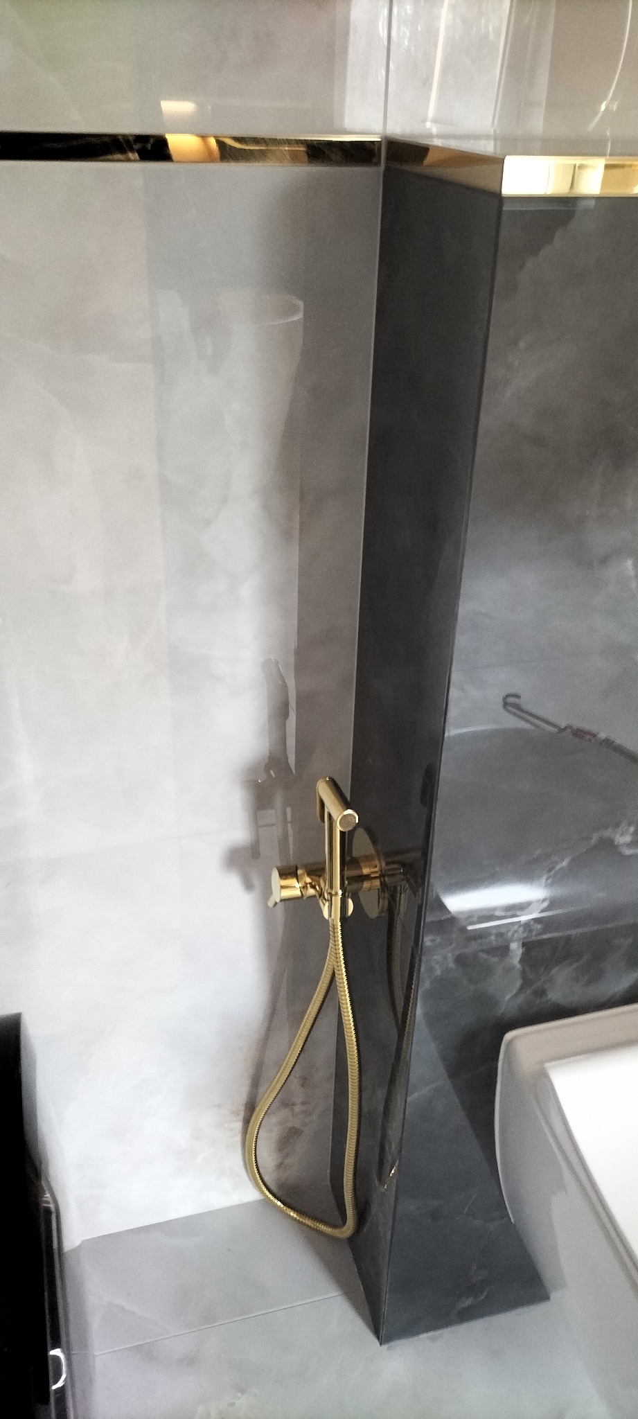 Błyszcząca, złota bateria prysznicowa na tle marmurowych płytek w odcieniach szarości i bieli, wykończonych złotą listwą. Nowoczesna aranżacja łazienki.