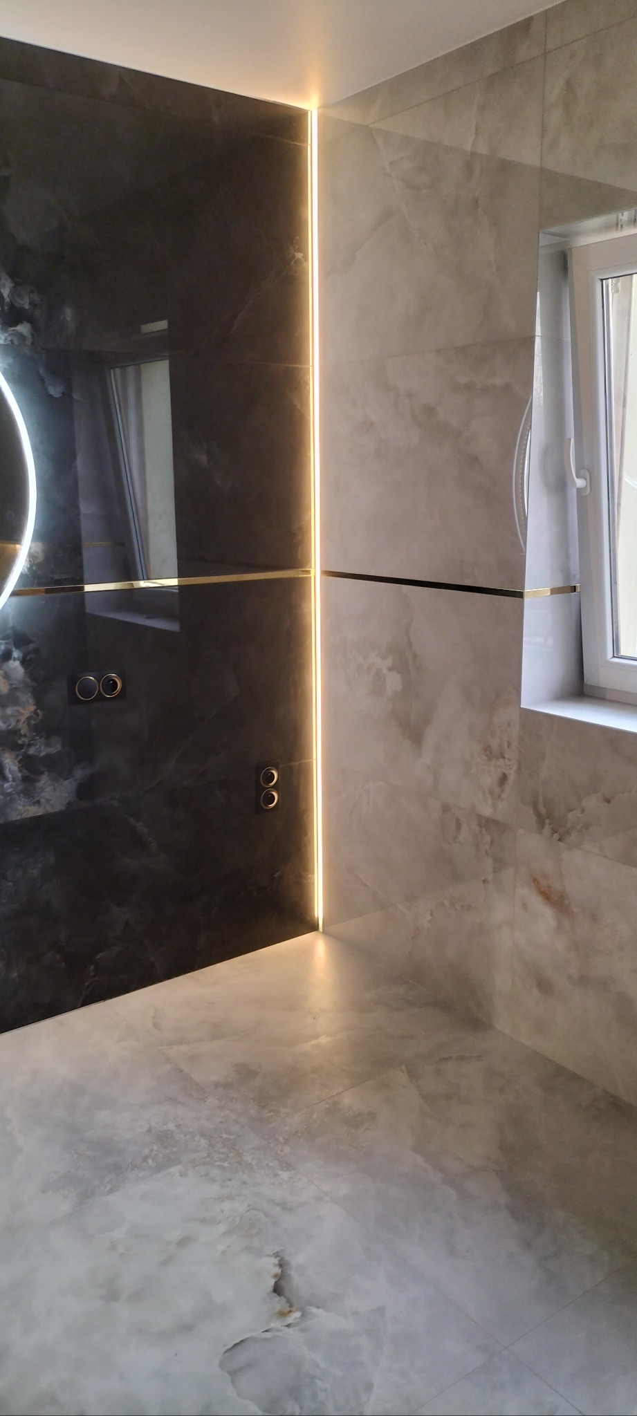 Nowoczesna łazienka z czarno-białymi płytkami imitującymi marmur, podświetleniem LED i złotymi akcentami. Widoczne gniazdka i fragment okrągłego lustra.