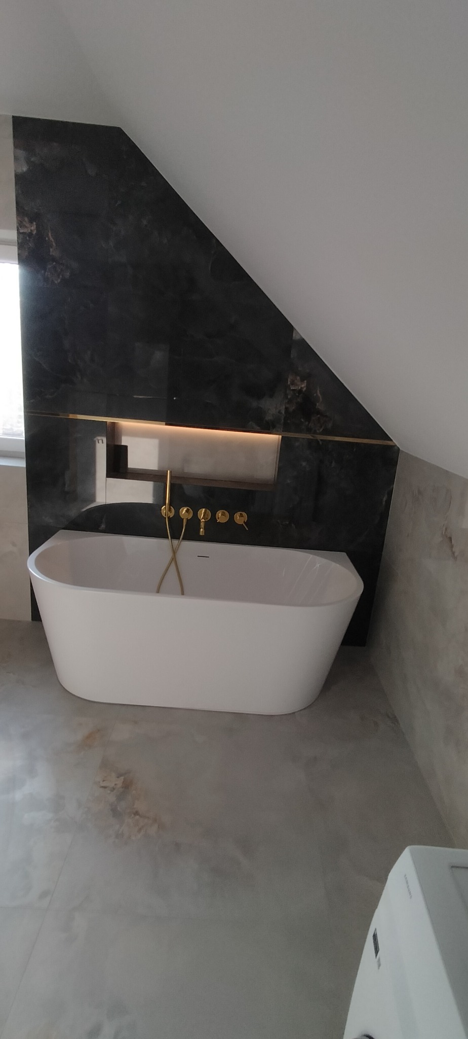 Nowoczesna łazienka na poddaszu z wolnostojącą wanną, czarnymi płytkami imitującymi marmur i złotą armaturą. Oświetlona wnęka nad wanną dodaje elegancji.