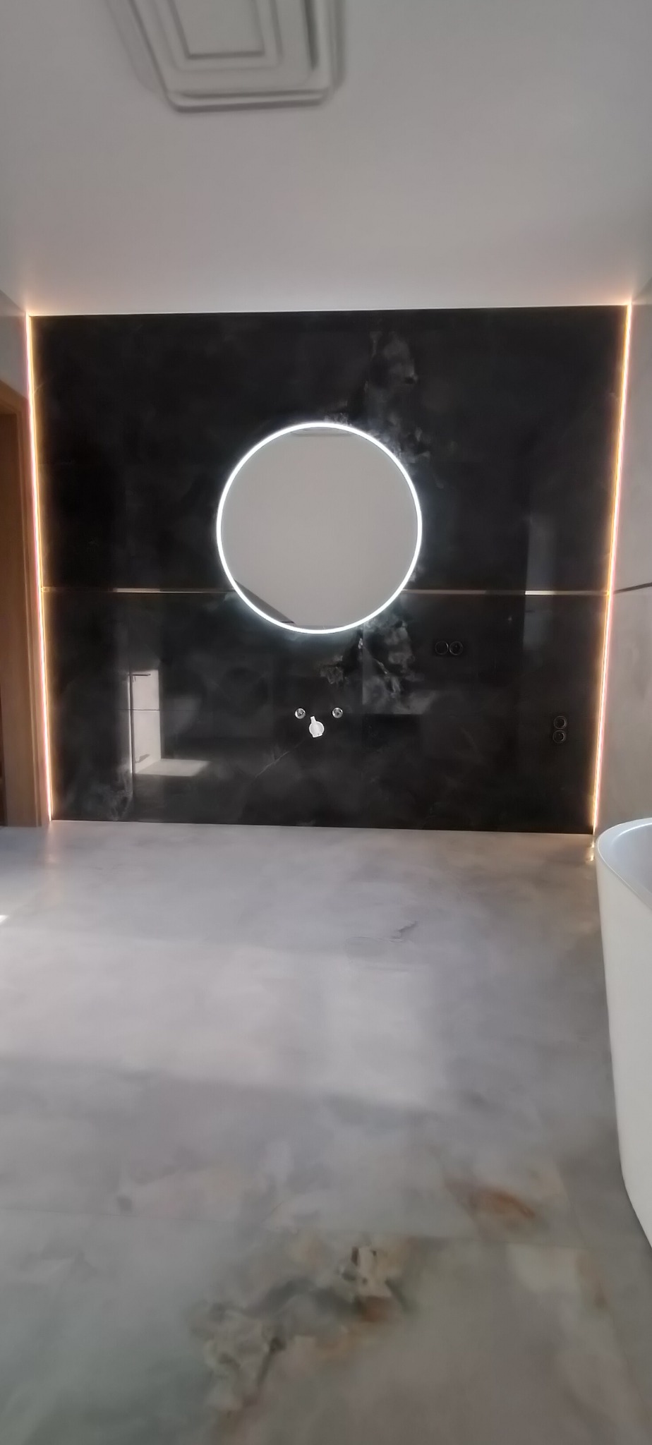 Nowoczesna łazienka z czarną ścianą imitującą marmur, okrągłym lustrem z podświetleniem LED i szarą posadzką żywiczną. Minimalistyczny design z akcentami świetlnymi.