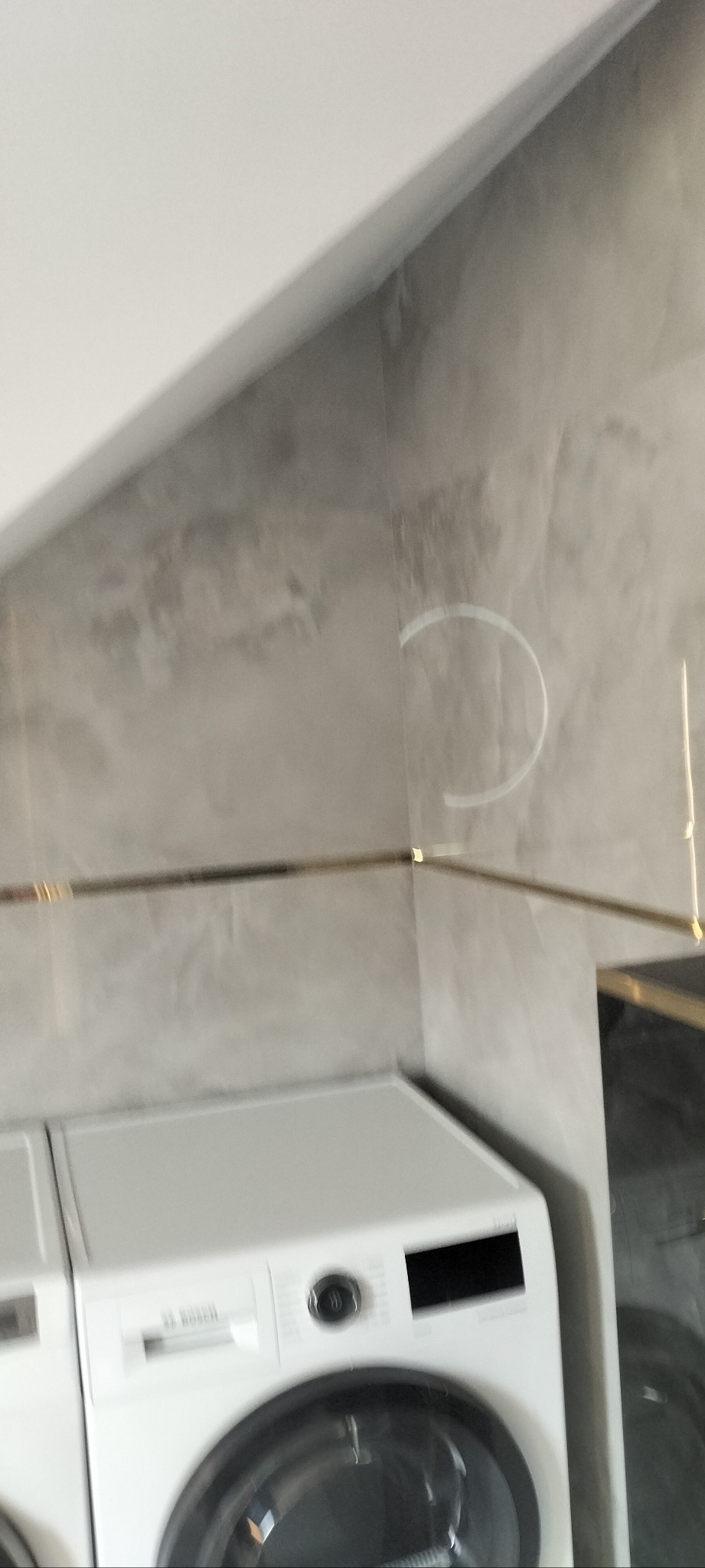 Szara ściana z efektem betonu w łazience, wykończona złotą listwą, z widoczną pralką Bosch. Minimalistyczny design, nowoczesne wykończenie.