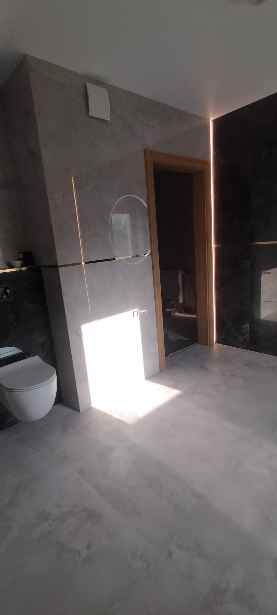 Nowoczesna łazienka z szarymi płytkami imitującymi marmur, podświetleniem LED i wiszącą toaletą. Widoczne okrągłe lustro i drewniane obramowanie drzwi.