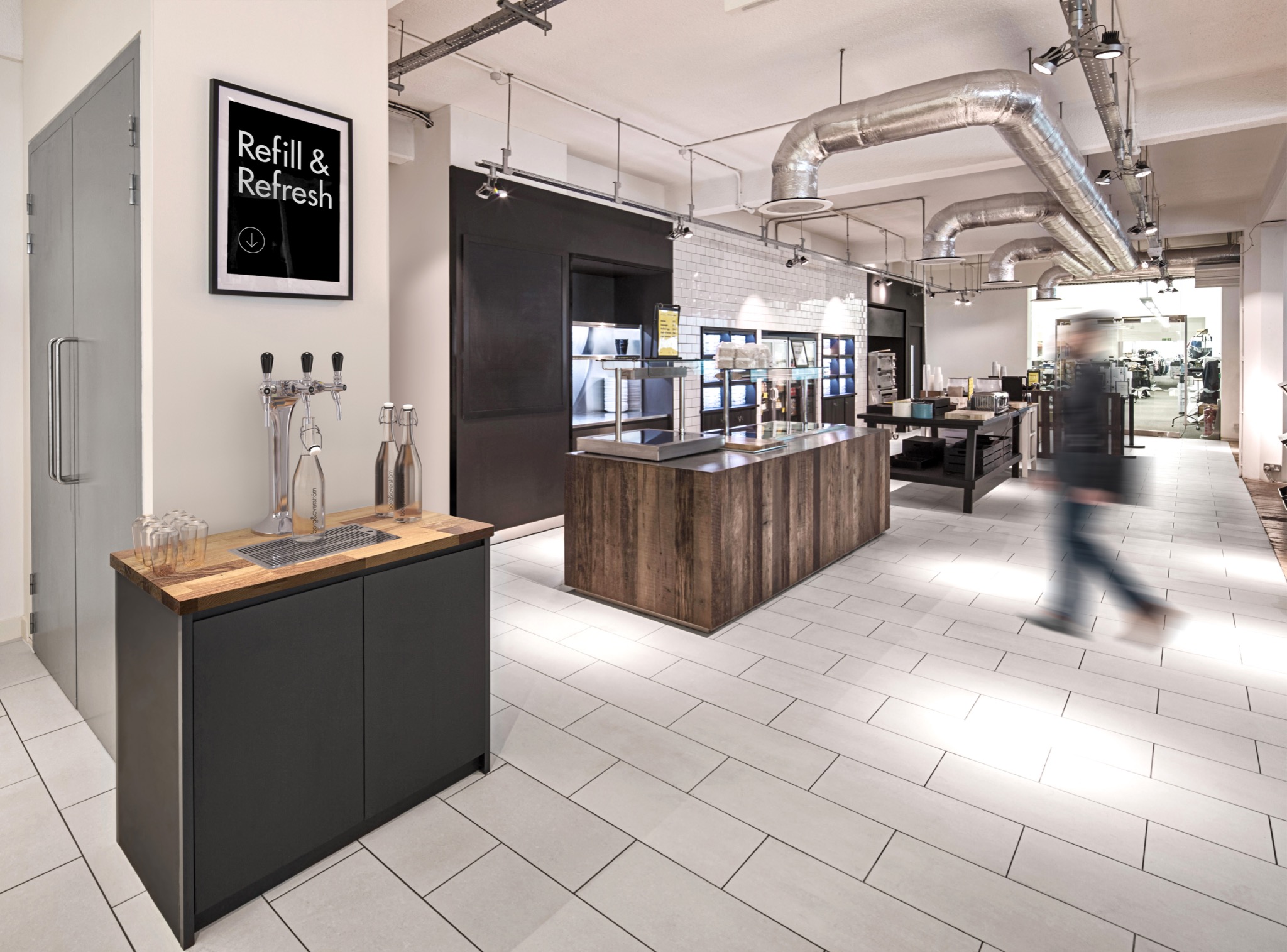 Wnętrze biurowe z dystrybutorem wody z kranami, butelkami i szklankami, plakat 'Refill & Refresh', widoczne instalacje wentylacyjne pod sufitem i osoba w ruchu w tle.