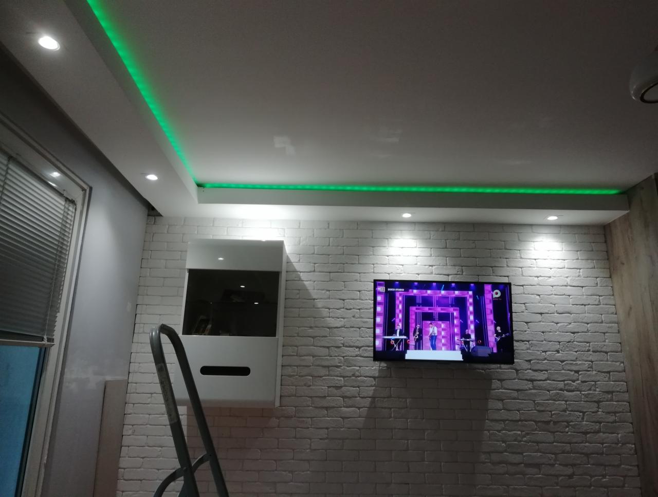 Pokój z białymi ceglanymi ścianami, podświetlanym zielonym LED sufitem, telewizorem i białą szafką wiszącą, widoczna drabina.