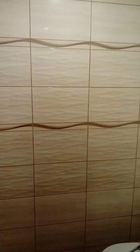 Ściana wyłożona beżowymi płytkami ceramicznymi z subtelnym, falistym wzorem i brązowym dekoracyjnym pasem, sfotografowana z bliska.