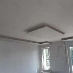 Montaż sufitu podwieszanego z płyt karton-gips. Widoczne płyty na konstrukcji nośnej, przygotowane do szpachlowania. Dwa okna w tle.