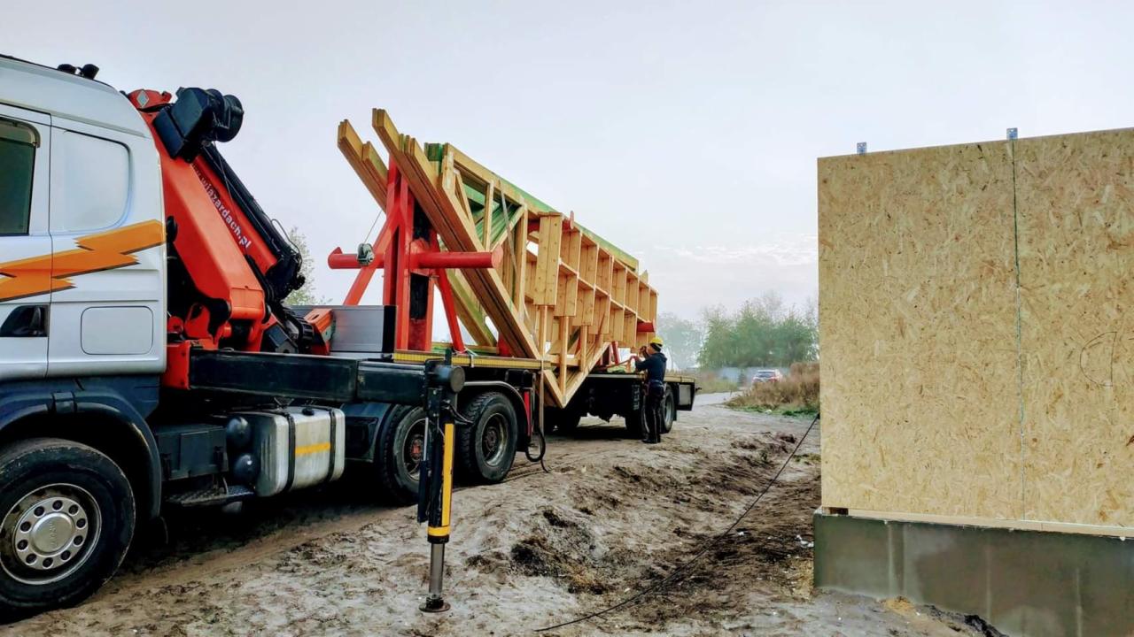 Ciężarówka z dźwigiem przewozi drewniane elementy konstrukcyjne domu szkieletowego na plac budowy, obok stoi już fragment ściany z płyty OSB.